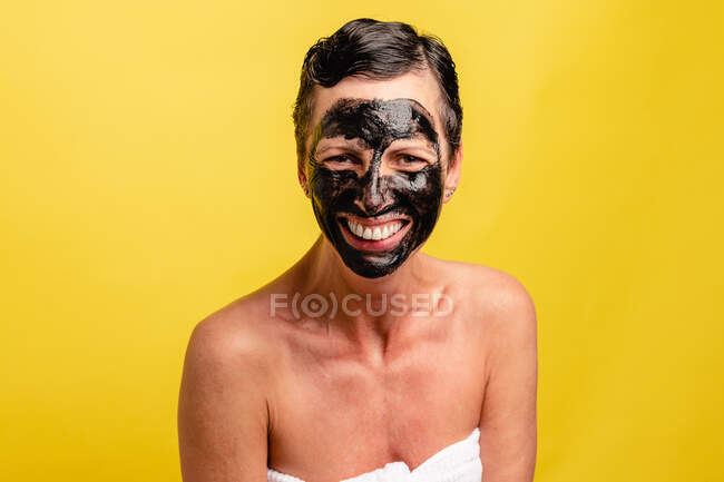 Allegro femmina di mezza età con peel off maschera nera in piedi su sfondo giallo studio alla ricerca di una fotocamera — Foto stock