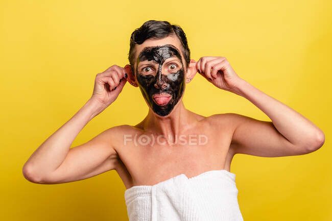 Mujer de mediana edad con máscara de piel negra en la cara mirando a la cámara con la lengua hacia fuera en el fondo amarillo - foto de stock