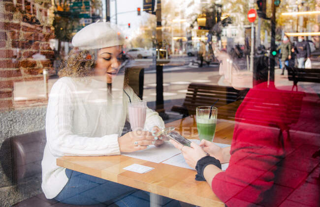 View through a glass of two friends looking at their cell phones inside a bar while having a fruit smoothie — Stock Photo