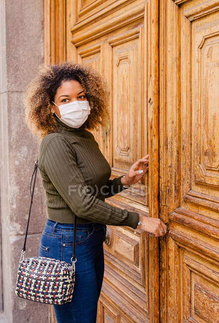 Afro donna con maschera che apre la porta di casa mentre guarda la macchina fotografica — Foto stock