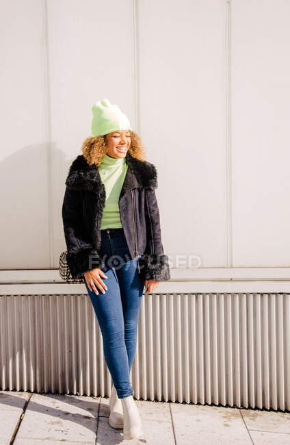 Corpo inteiro de uma mulher afro que está ao lado de uma parede na rua sorrindo em um dia ensolarado enquanto vestindo um casaco e chapéu — Fotografia de Stock