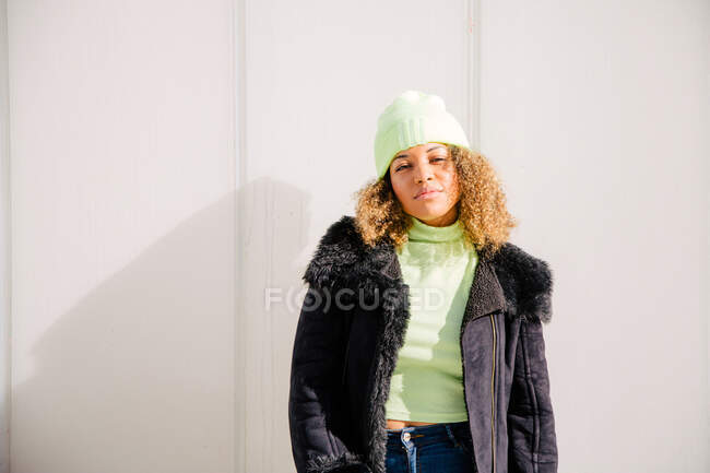 Afro femme qui est debout près d'un mur dans la rue par une journée ensoleillée tout en portant une veste et un chapeau et en regardant la caméra — Photo de stock