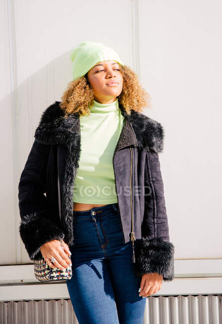 Afro donna che è in piedi da un muro in strada in una giornata di sole mentre indossa una giacca e cappello e guardando altrove — Foto stock