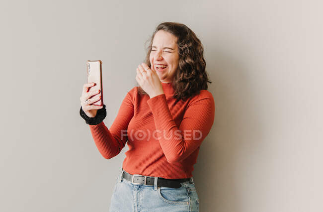 Mujer haciendo una videollamada mientras ríe junto a una pared blanca - foto de stock