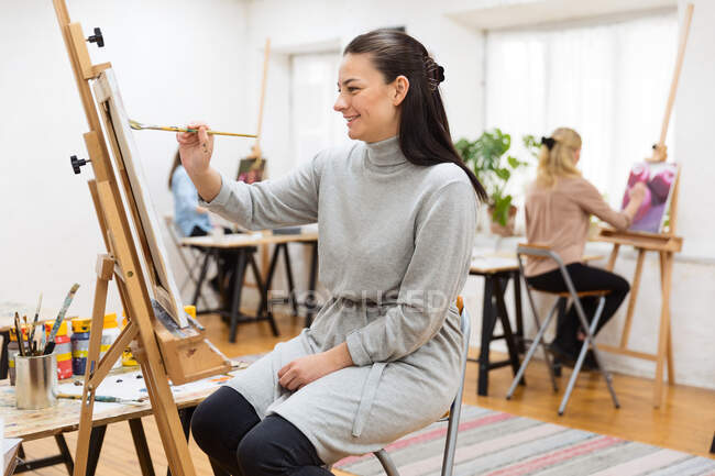 Vista laterale della pittura focalizzata artista femminile su tela su cavalletto in studio d'arte su sfondo di donne sfocate — Foto stock