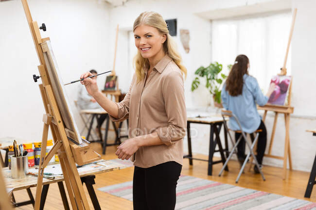 Vista lateral de la artista femenina encantada mirando a la cámara mientras pinta sobre lienzo sobre caballete en estudio de arte sobre fondo de mujeres borrosas - foto de stock