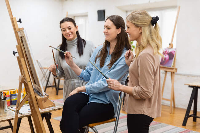 Groupe d'artistes féminines heureuses se réunissant autour du chevalet avec la peinture pendant l'atelier dans l'atelier d'art — Photo de stock