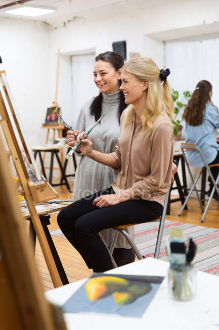 Vista lateral do artista feminino ensinar mulher pintura quadro no cavalete durante oficina no estúdio criativo — Fotografia de Stock
