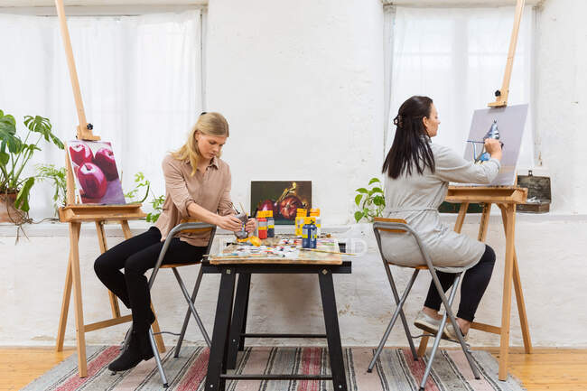 Vista lateral da pintura artista feminina focada na tela no cavalete no estúdio de arte no fundo de mulheres desfocadas — Fotografia de Stock