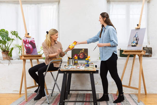 Вид сбоку улыбающейся женщины, передающей краску подруге во время рисования в художественной мастерской — стоковое фото