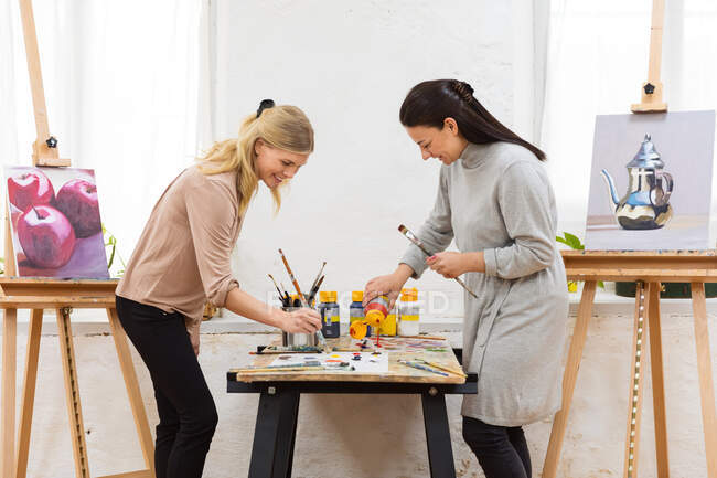 Обзор положительных женщин-художников, стоящих за столом с красками и создающих произведения искусства в мастерской — стоковое фото
