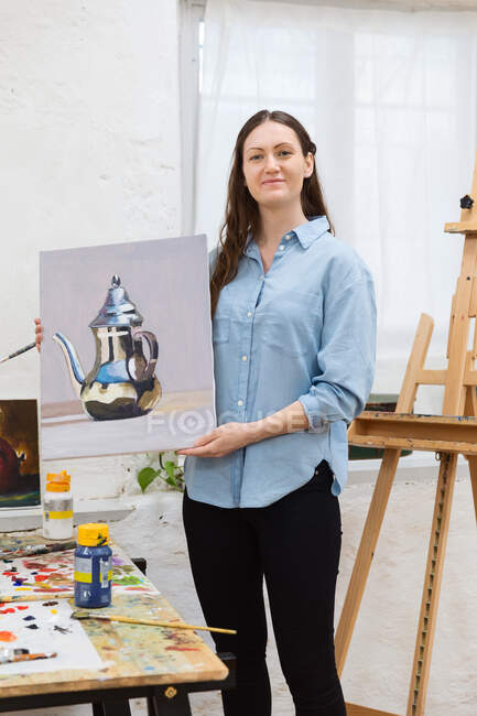 Artista femenina encantada de pie con pintura sobre lienzo en taller creativo y mirando a la cámara - foto de stock