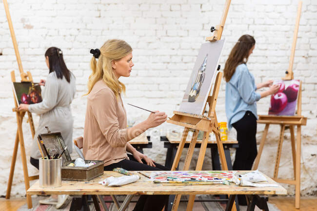 Vista lateral de la pintura de artista femenina enfocada en lienzo sobre caballete en estudio de arte sobre fondo de mujeres borrosas - foto de stock