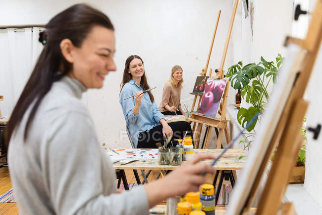 Vue latérale de la compagnie des amies heureuses peignant sur des toiles pendant l'atelier dans le studio d'art — Photo de stock