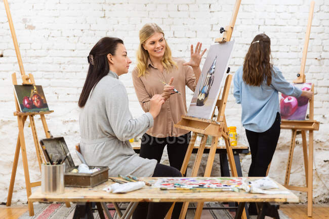 Vista lateral de la compañía de amigas felices pintando sobre lienzos durante el taller en el estudio de arte - foto de stock
