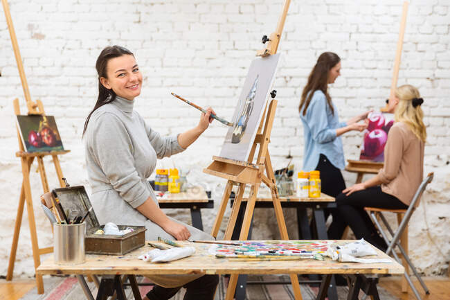 Vista laterale di artista femminile felice guardando la fotocamera mentre dipinge su tela su cavalletto in studio d'arte su sfondo di donne sfocate — Foto stock