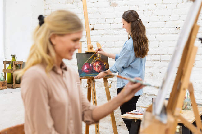 Vista lateral de la pintura artista femenina encantada sobre lienzo sobre caballete en estudio de arte sobre fondo de mujeres borrosas - foto de stock
