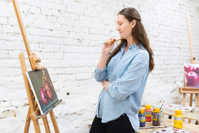 Side view of pensive female painter with brush standing near easel and looking at canvas with painting in art workshop — Stock Photo