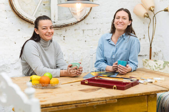 Artiste sorridenti sedute a tavola con pittura e vernici mentre bevono bevande nel laboratorio creativo e guardano la macchina fotografica — Foto stock