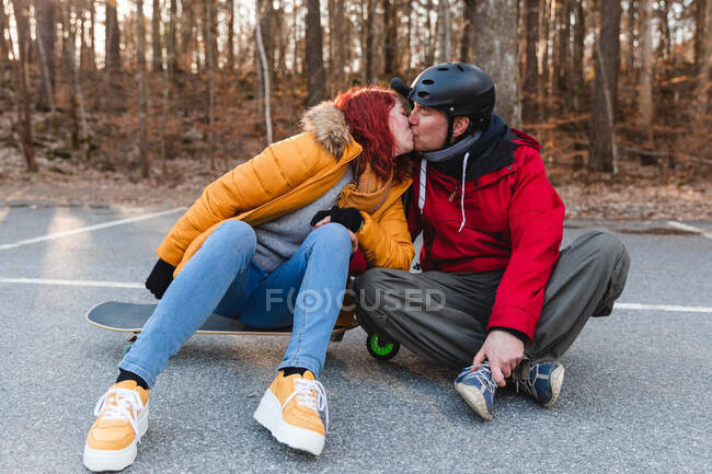 Encantada pareja sentada en monopatín y scooter mientras se besa y se divierte en el estacionamiento en otoño - foto de stock