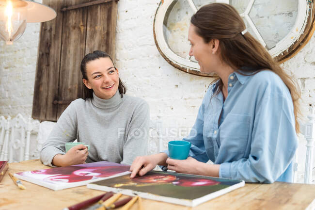 Весела жінка-художниця п'є напої під час обговорення живопису за столом в художній майстерні — стокове фото