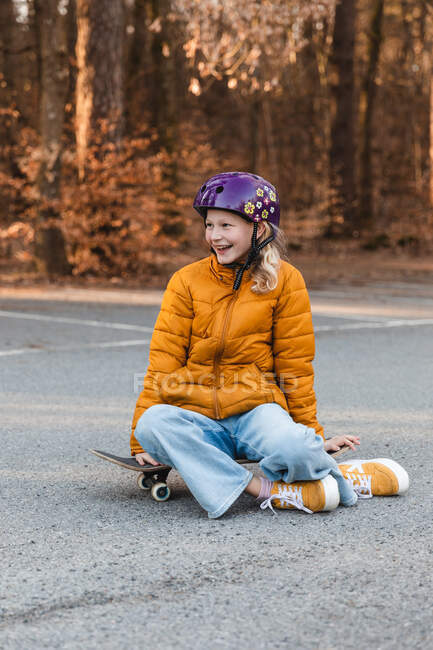 Sonriente chica adolescente en el casco sentado en el monopatín en el estacionamiento mientras disfruta de fin de semana en otoño y mirando hacia otro lado - foto de stock