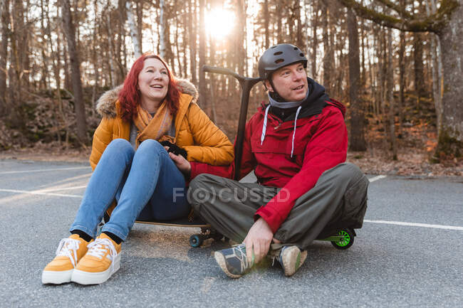 Encantada pareja sentada en monopatín y scooter mientras se divierten en el estacionamiento en otoño - foto de stock