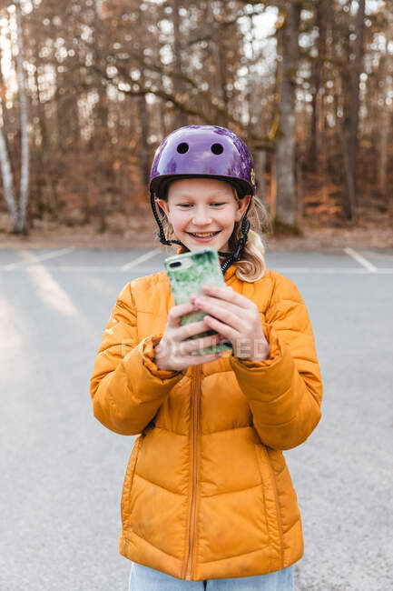 Fröhliches Teenager-Mädchen mit Helm fotografiert mit dem Handy auf dem Parkplatz im Herbstpark und blickt in die Kamera — Stockfoto