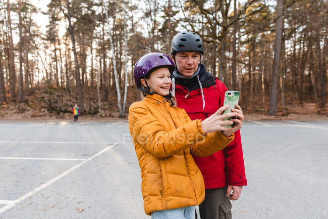 Fröhlicher Vater und Teenager mit Helm stehen am Herbstwochenende auf dem Parkplatz im Park und machen Selbstporträt mit dem Handy — Stockfoto
