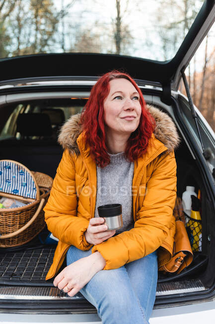 Спокойная женщина в осеннем пальто сидит в багажнике автомобиля и пьет горячий чай из термоса чашки, наслаждаясь дорожной поездкой и глядя в сторону — стоковое фото