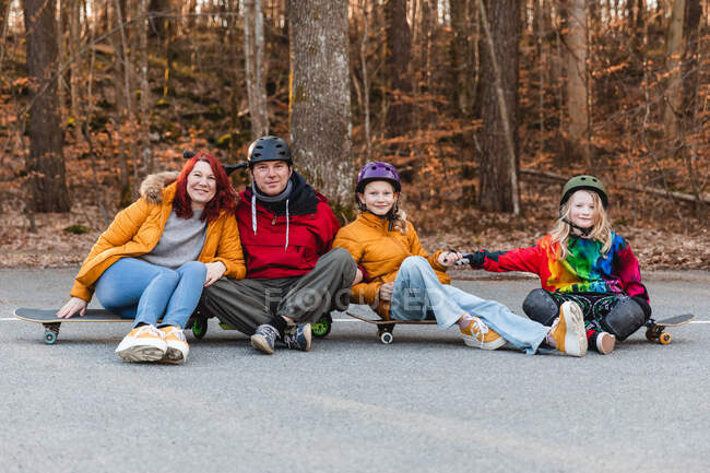 Positive Familie mit Helm sitzt auf Skateboards auf dem Parkplatz und blickt in die Kamera, während sie am Wochenende im Herbstpark Spaß hat — Stockfoto