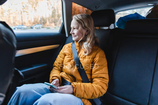 Contenido chica adolescente sentada en el coche en el asiento del pasajero y la navegación del teléfono móvil mientras mira por la ventana - foto de stock