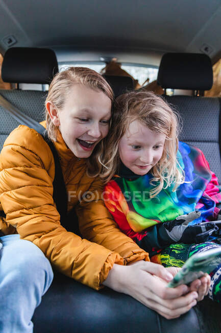 Возбужденные сестры сидят в машине и вместе играют в интересную игру на мобильном телефоне — стоковое фото