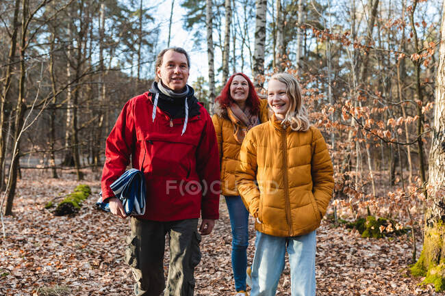 Sonriente pareja e hija adolescente caminando con cuadros en el bosque para hacer un picnic en otoño - foto de stock
