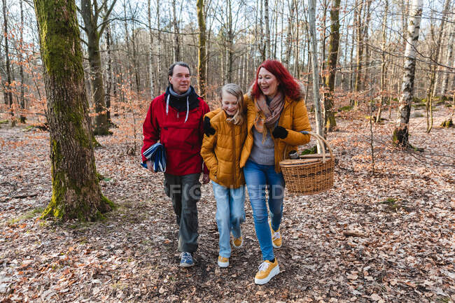 Pareja sonriente e hija adolescente caminando con cesta y cuadros en el bosque para hacer un picnic en otoño - foto de stock