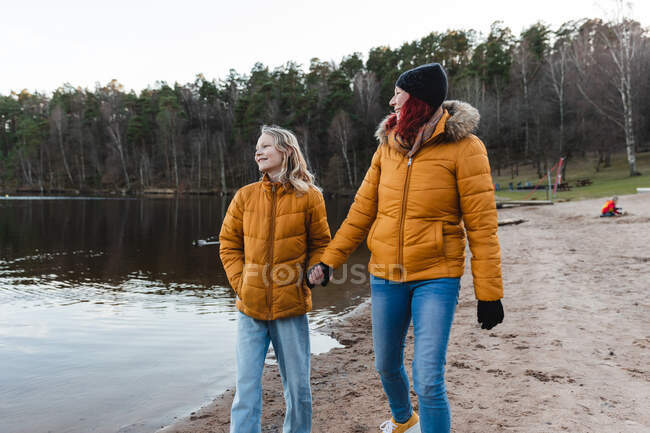 Madre contenta y chica adolescente tomados de la mano y caminando por el estanque en el bosque mientras disfruta del fin de semana en otoño - foto de stock