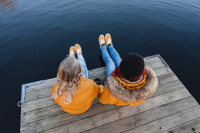 Зверху невизначена мати і дівчинка-підліток, що сидить на дерев'яній набережній біля ставка разом восени — стокове фото