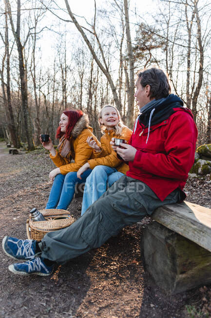 Seitenansicht einer glücklichen Familie, die Tee aus Thermoskannen trinkt und Essen isst, während sie im Herbst im Wald picknickt — Stockfoto