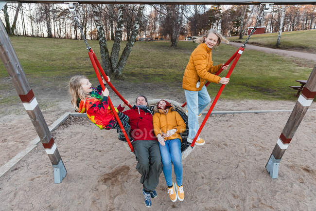 Desde arriba de la familia encantada montar columpio red en el parque infantil en el otoño y divertirse juntos - foto de stock