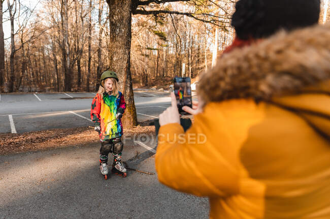 Невідома мати фотографує усміхнену доньку в шоломі і роликові лопаті в осінньому парку — стокове фото