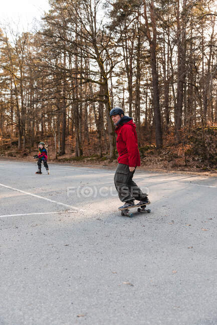 Отец в шлеме катается на скейтборде и ребенок катается на роликах на парковке, веселясь вместе в осеннем парке — стоковое фото