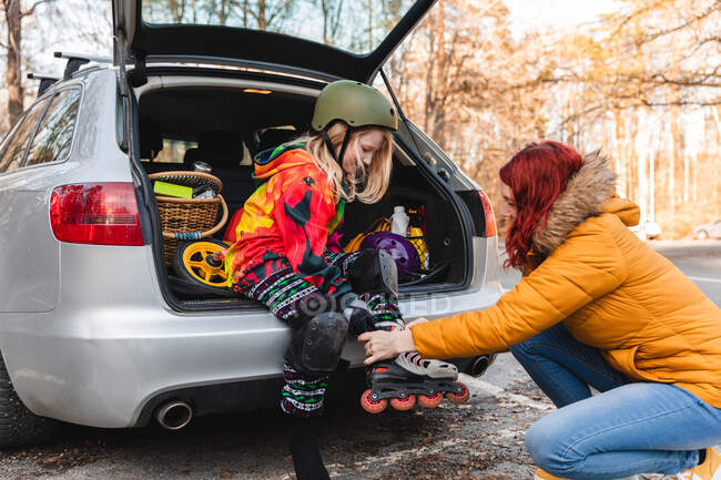 Vista lateral de la madre ayudando a la niña a ponerse patines mientras está sentada en el maletero del coche - foto de stock