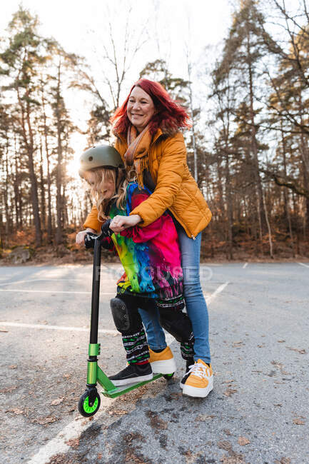 Delighted mother and daughter riding kick scooter on parking while having fun at weekend in autumn — Stock Photo