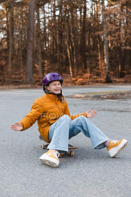 Cheerful teen girl in helmet sitting on skateboard in autumn park and having fun during weekend — Stock Photo