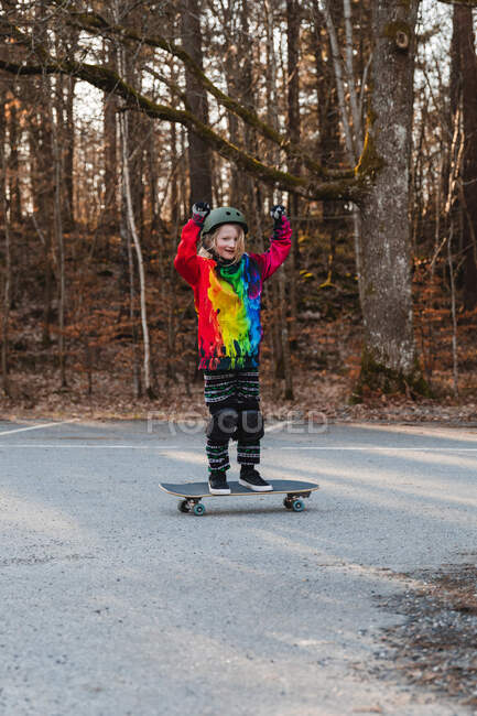 Счастливая девушка в защитном шлеме стоит на скейтборде и празднует победу с поднятыми руками, веселясь в осеннем парке — стоковое фото