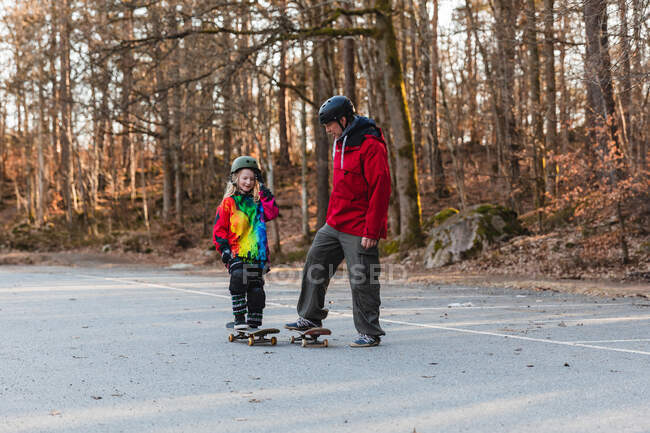 Padre e figlia spensierati nei caschi cavalcando skateboard nel parco e divertendosi insieme durante il fine settimana — Foto stock