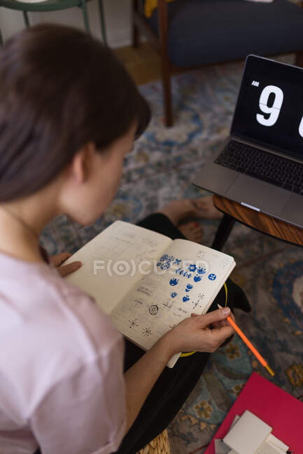 Disegno grafico femminile focalizzato in sketchbook mentre si lavora a distanza sul progetto a tavola con una tazza di caffè a casa — Foto stock