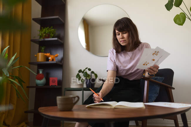 Full body barefoot woman sitting near table and examining paper sheets with sketches while working on creative project at home — Stock Photo