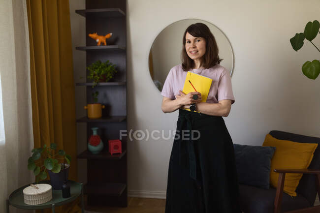 Щаслива жінка з ескізом і олівцем посміхається і дивиться на камеру, стоячи біля крісла в творчій студії вдома — стокове фото