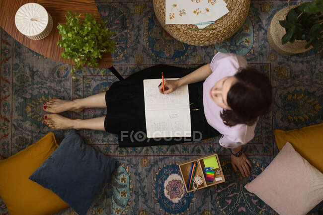 Artista femminile adulta a corpo pieno seduta al piano vicino al tavolo con laptop e schizzo di disegno in sketchbook a casa — Foto stock